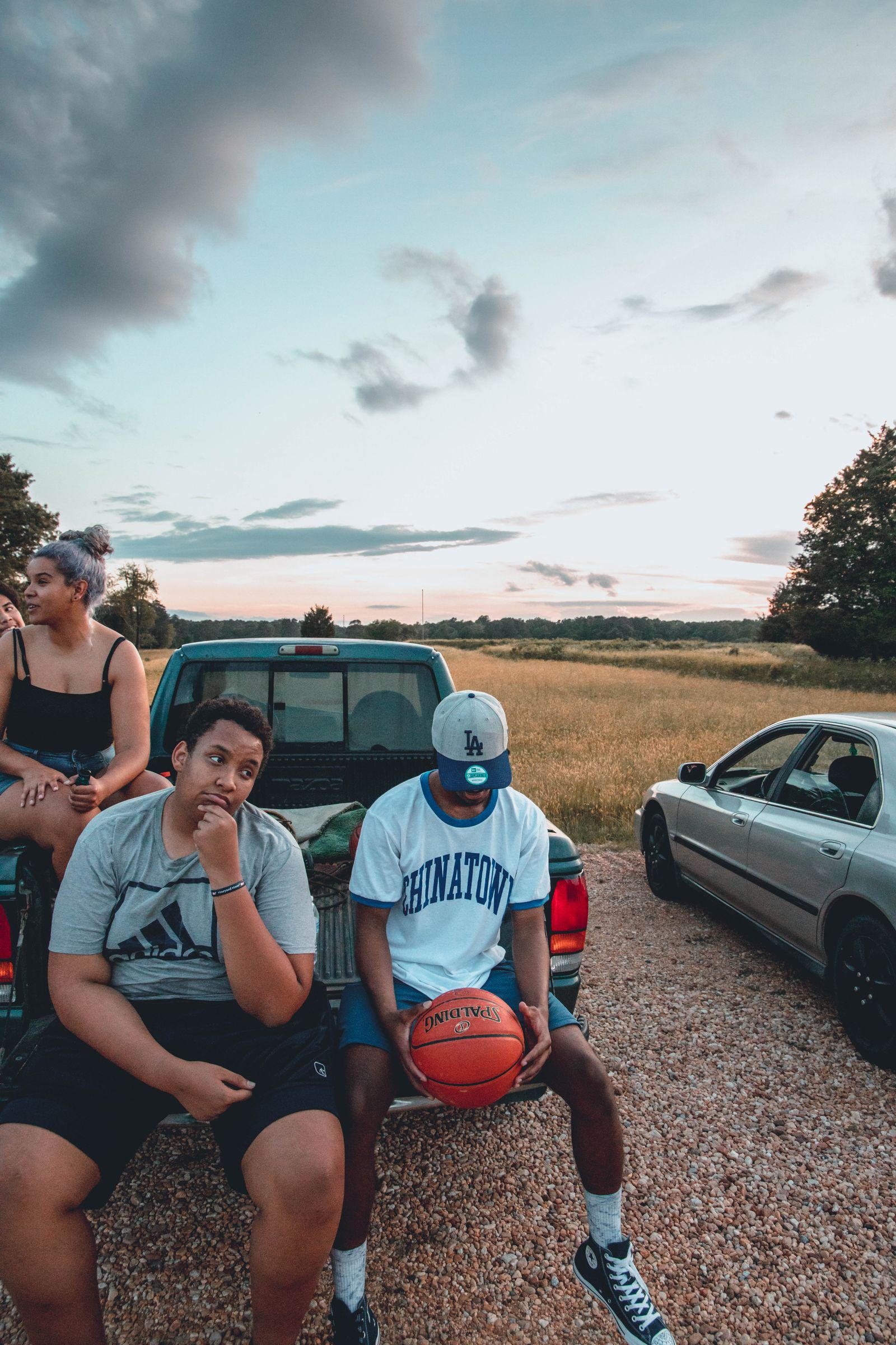 High angle of young friends sitting on car with basketball in parking lot while spending time together in nature